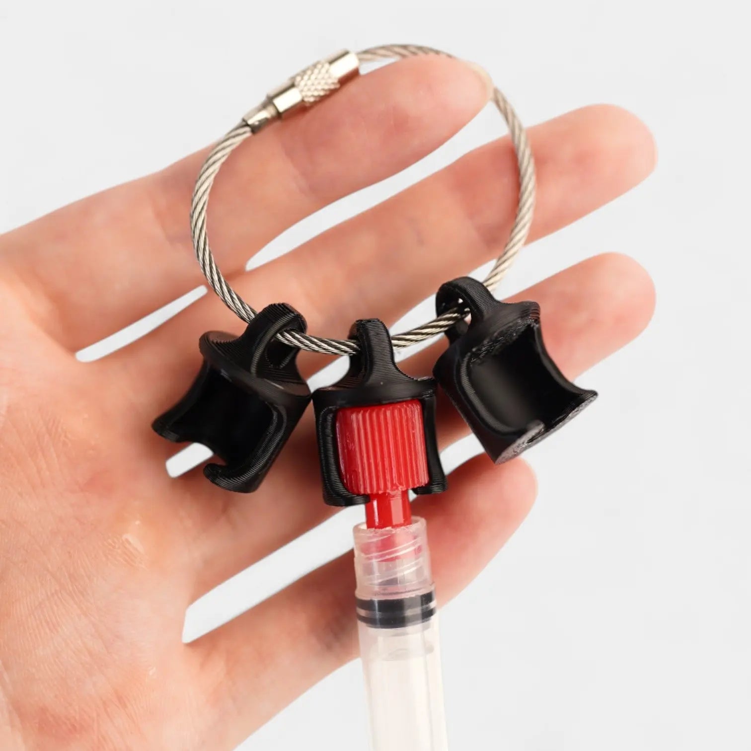Black polylactic acid created using additive manufacturing B-Braun cap clip on steel cable keyring. Featured attached to a syringe. Shown being held with a white background.