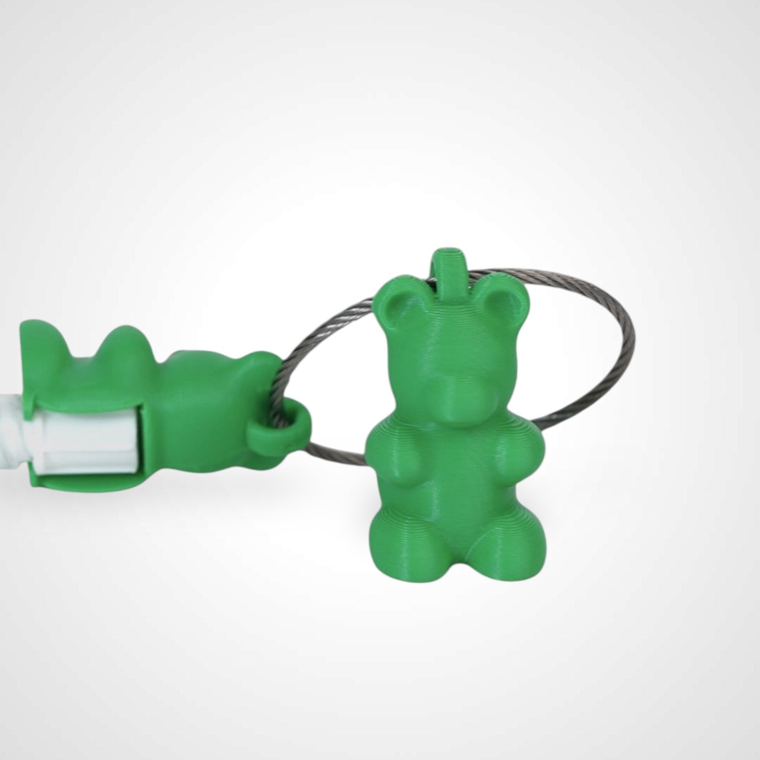 Two green polylactic acid created using additive manufacturing gummy bear cap clips on steel cable keyring. Featured attached to a syringe. Shown on a white background.