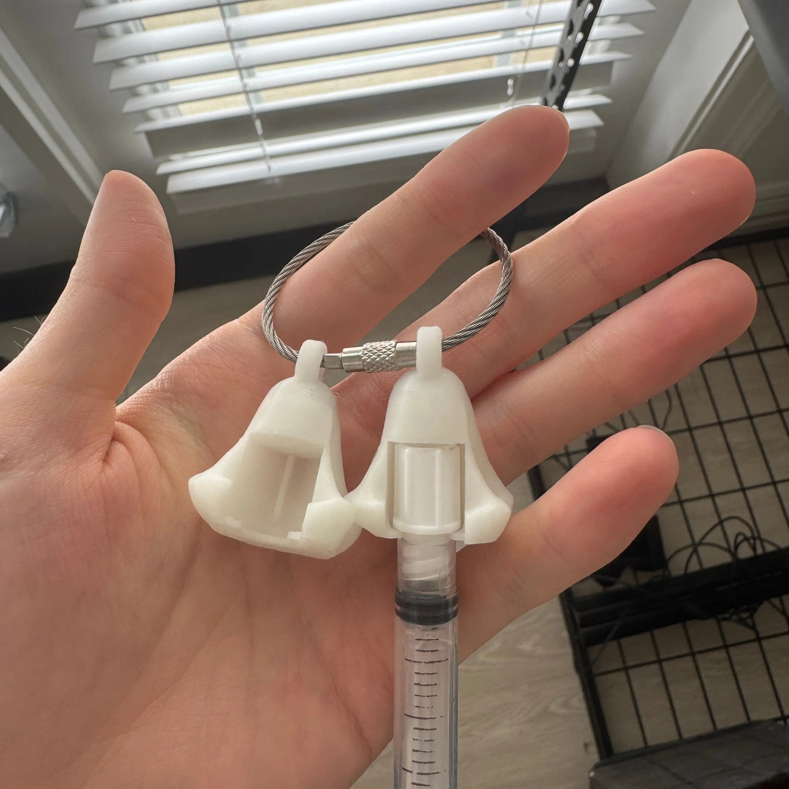 Two white polylactic acid created using additive manufacturing ghost cap clips on steel cable keyring. Featured attached to a syringe. Shown being held.