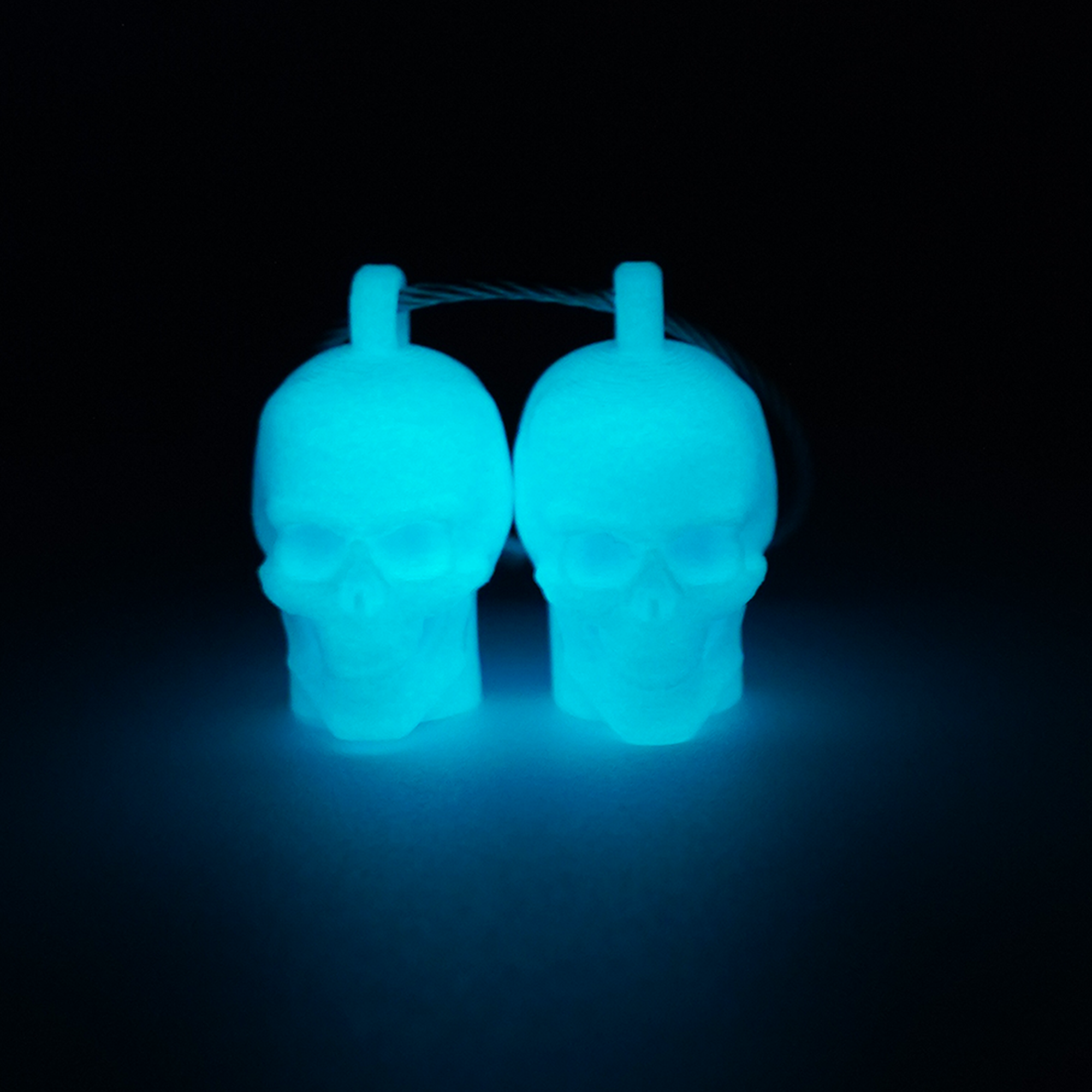 Two blue polylactic acid created using additive manufacturing skull cap clips on steel cable keyring. Shown glowing blue on a black background.