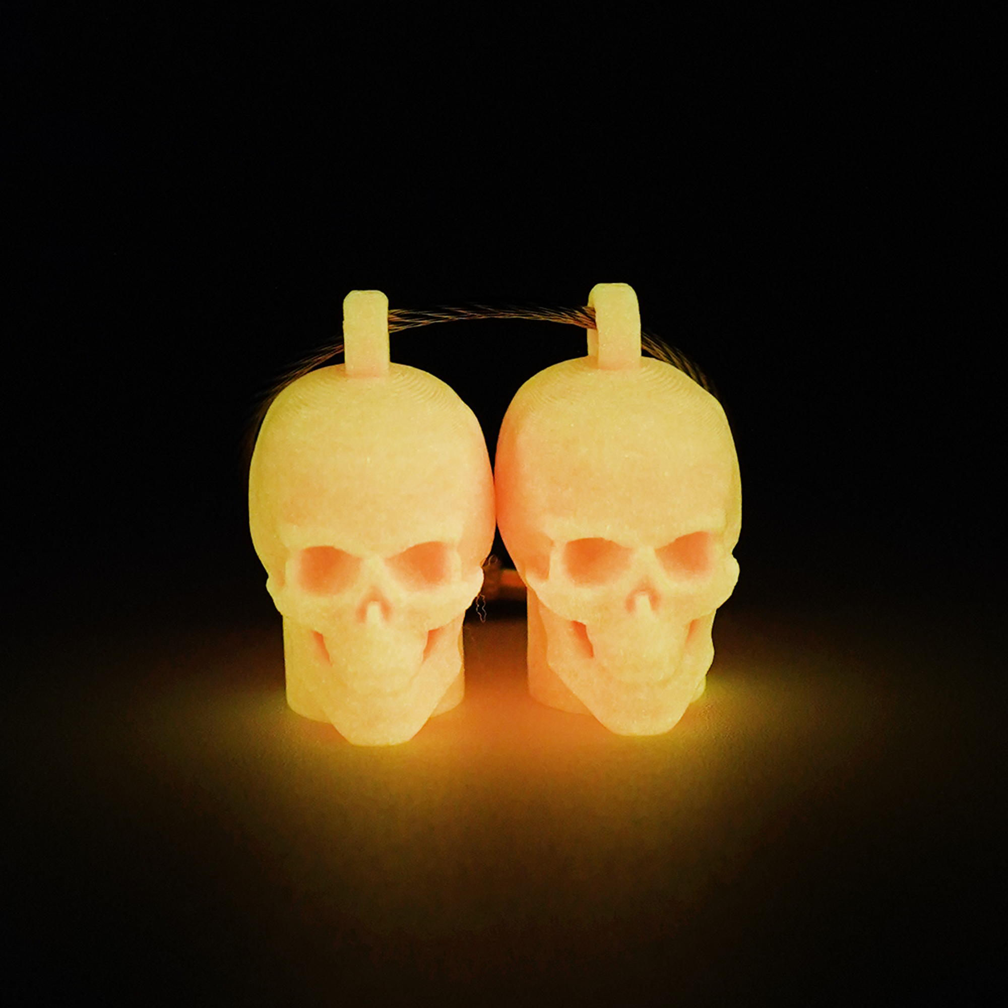 Two pink polylactic acid created using additive manufacturing skull cap clips on steel cable keyring. Shown glowing orange on a black background.