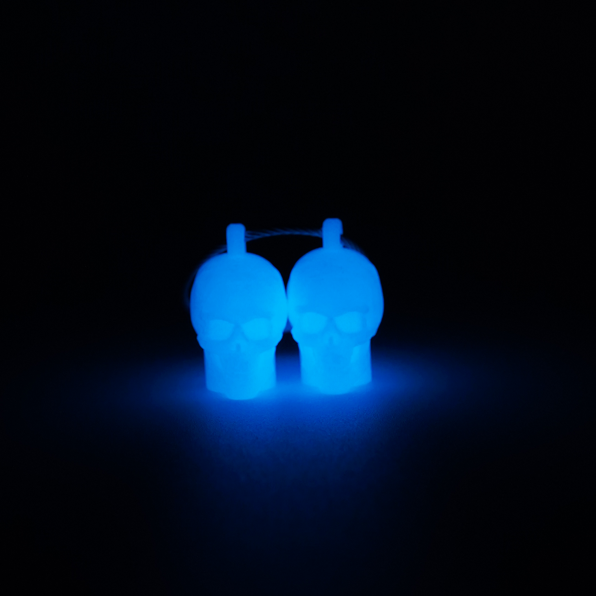Two white polylactic acid created using additive manufacturing skull cap clips on steel cable keyring. Shown glowing blue on black background.