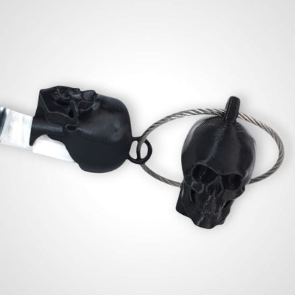 Two black polylactic acid created using additive manufacturing skull cap clips on steel cable keyring. Featured attached to a syringe. Shown on a white background.