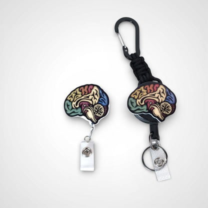 Sagittal brain badge reel. On a carabiner with a steel retractable cable next to a swivel-alligator clip that rotates 360° with a nylon retractable string. Shown on a white background. 