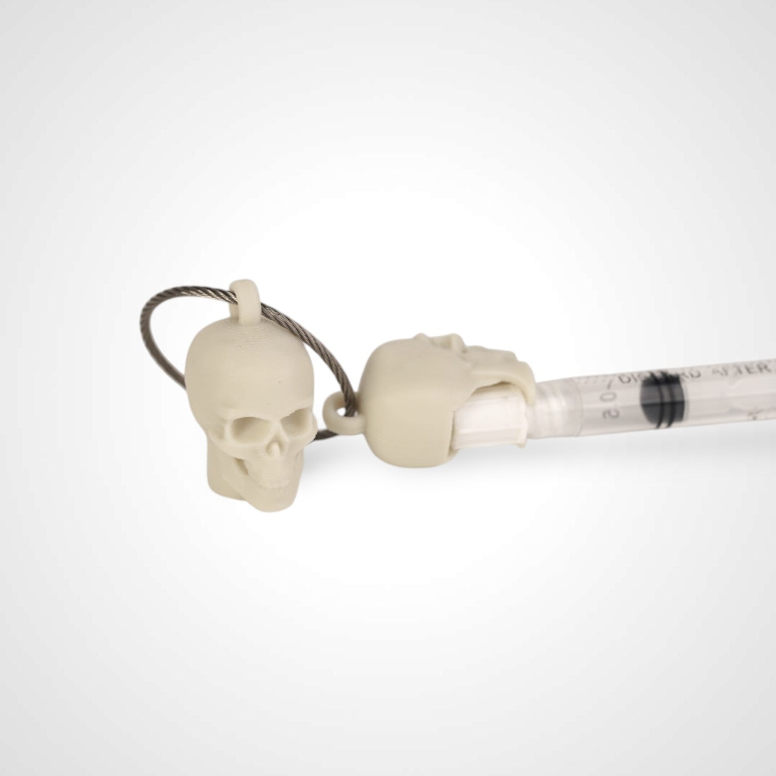 Two off white polylactic acid created using additive manufacturing skull cap clips on steel cable keyring. Featured attached to a syringe. Shown on a white background.