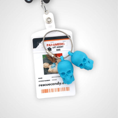 Two blue polylactic acid created using additive manufacturing skull cap clips on steel cable keyring. Featured attached to a badge. Shown on a white background.
