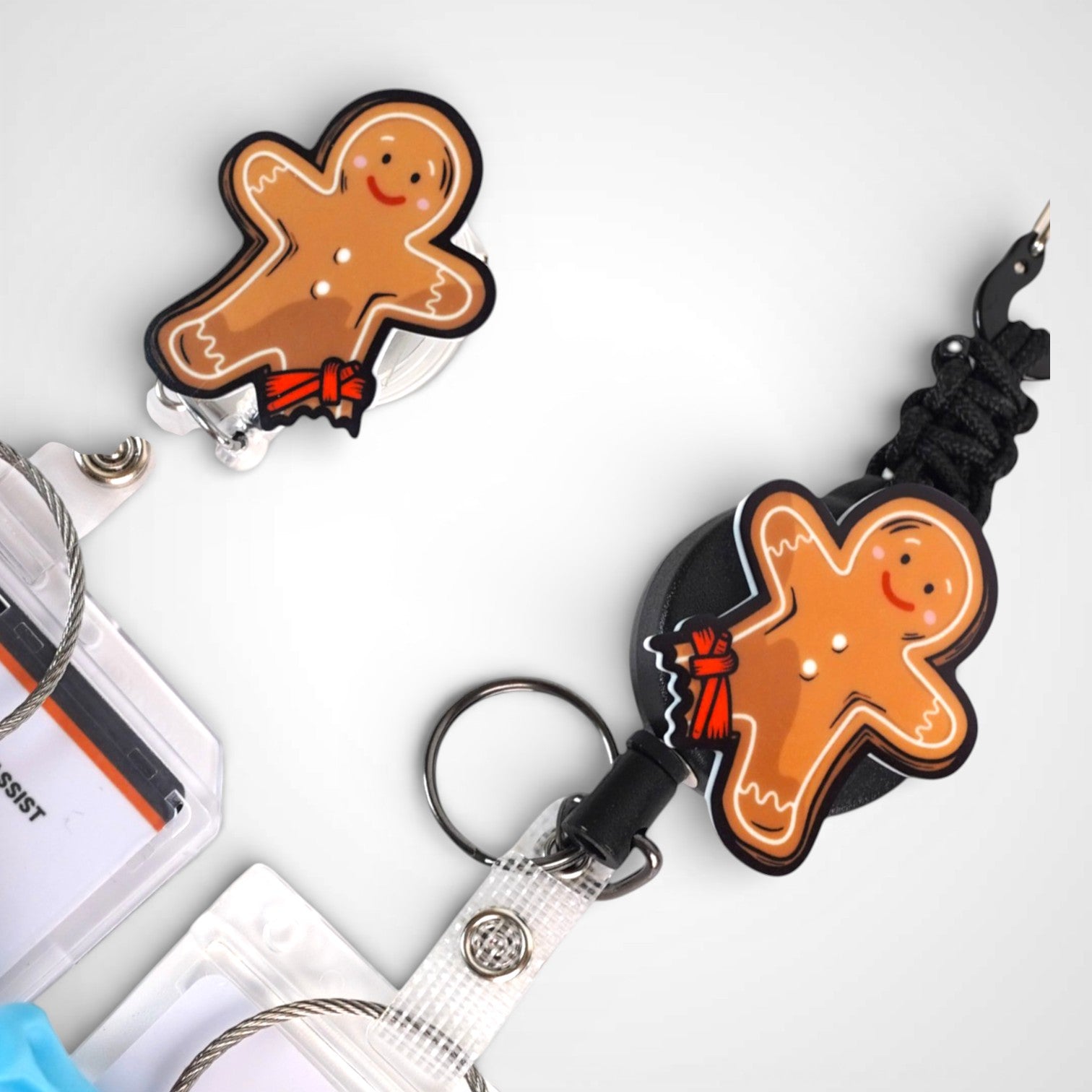 Tourniquet gingerbread man badge reel. On a carabiner with a steel retractable cable next to a swivel-alligator clip that rotates 360° with a nylon retractable string. Shown on a white background. 